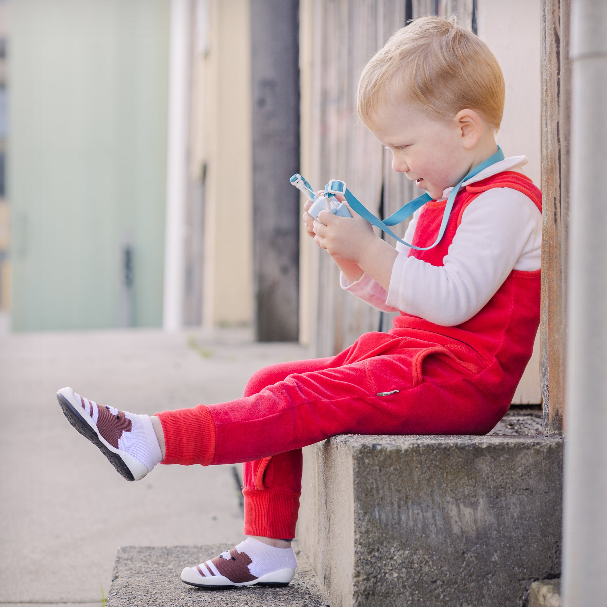 Blond toddler in red overalls, white shirt, and brown and white shoes sits on concrete steps, playing with a toy camera.