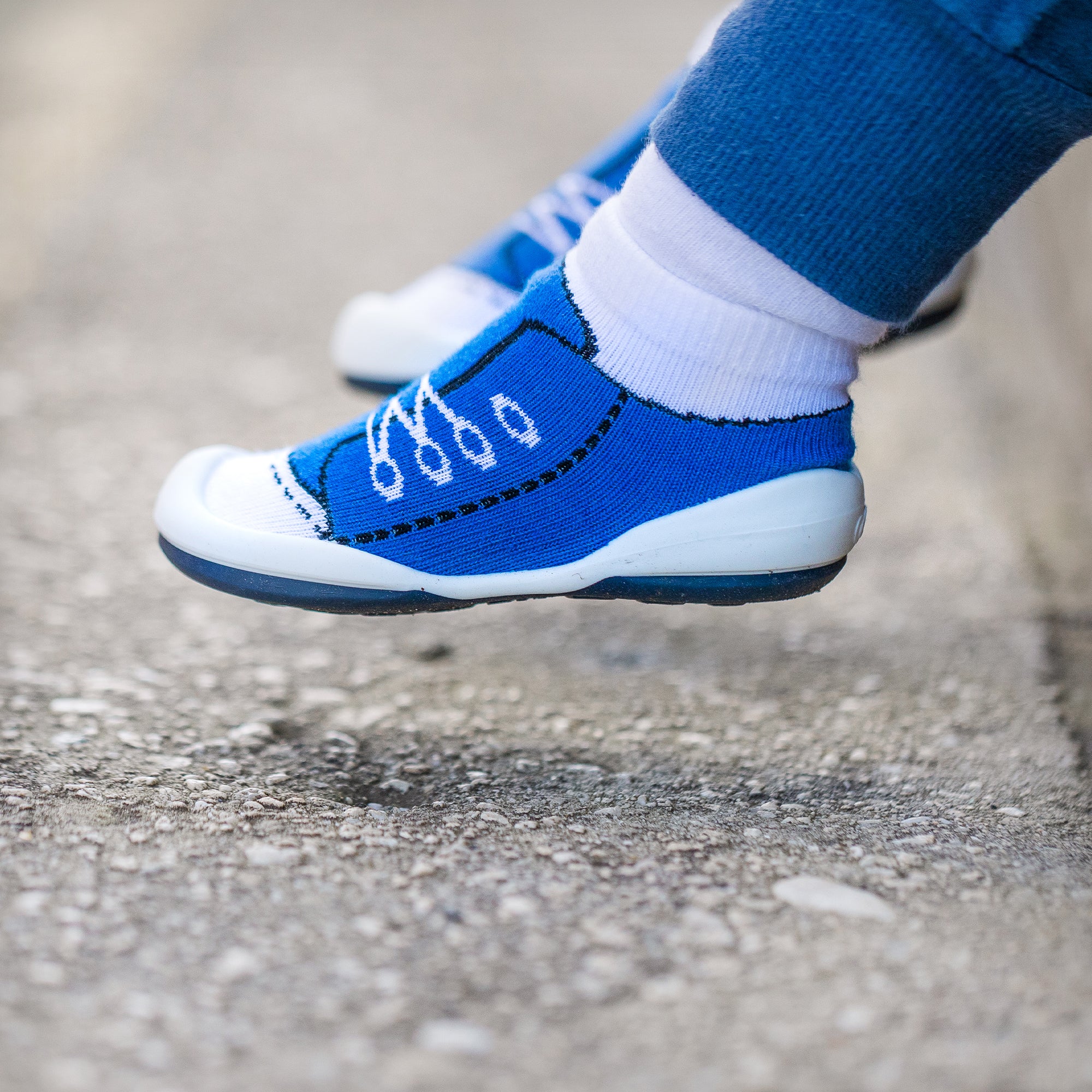 A child's blue sock shoes with a white toe cap and white shoelace design, paired with blue pants.