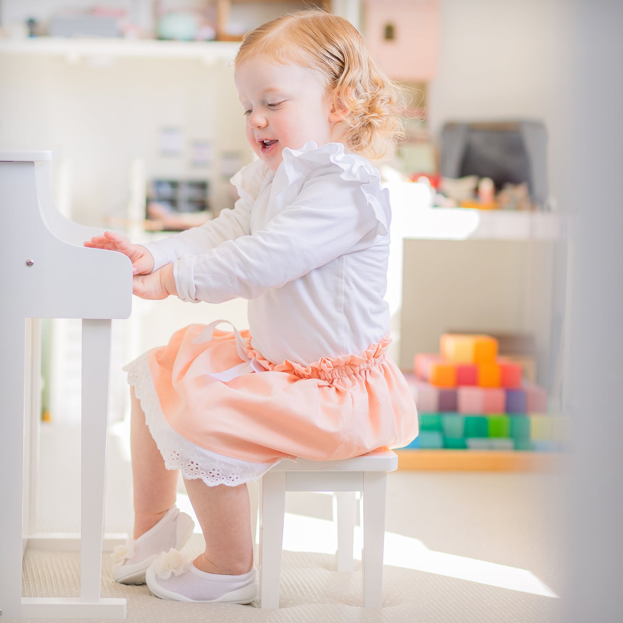 A toddler plays a white toy piano while wearing a white top, peach skirt, and white shoes.