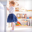 Young child with red hair, white long-sleeved shirt, denim skirt, and white shoes holding a small toy. Bright, organized playroom with shelves of toys.