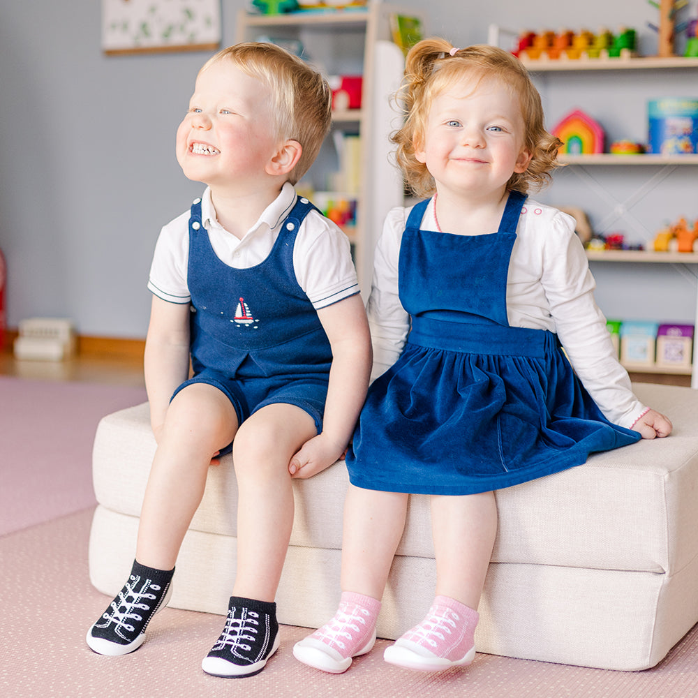 Two young children in blue outfits and shoe socks sitting on a white ottoman.