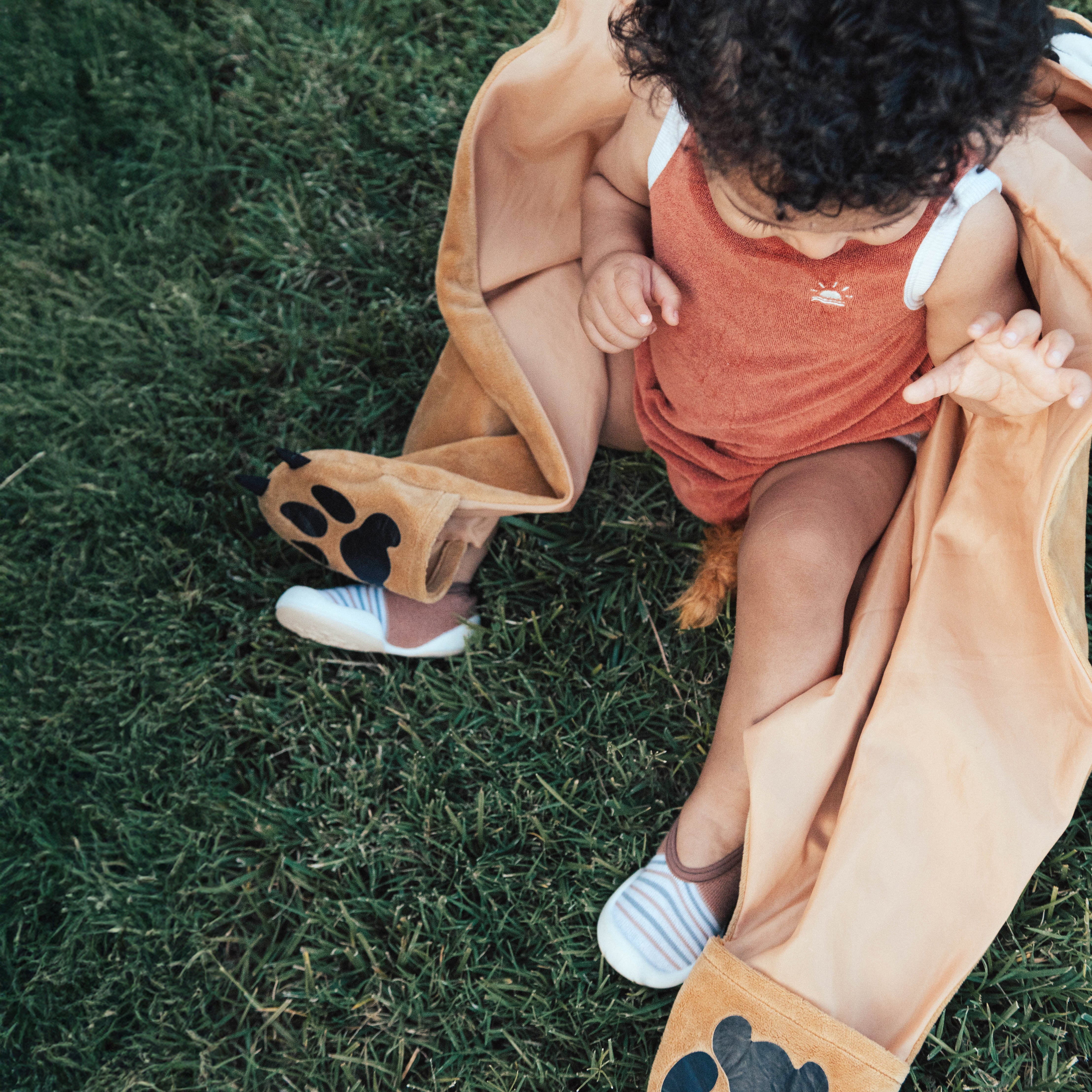 A baby with curly hair sits on green grass while wearing an orange romper, striped shoes, and a brown lion costume.