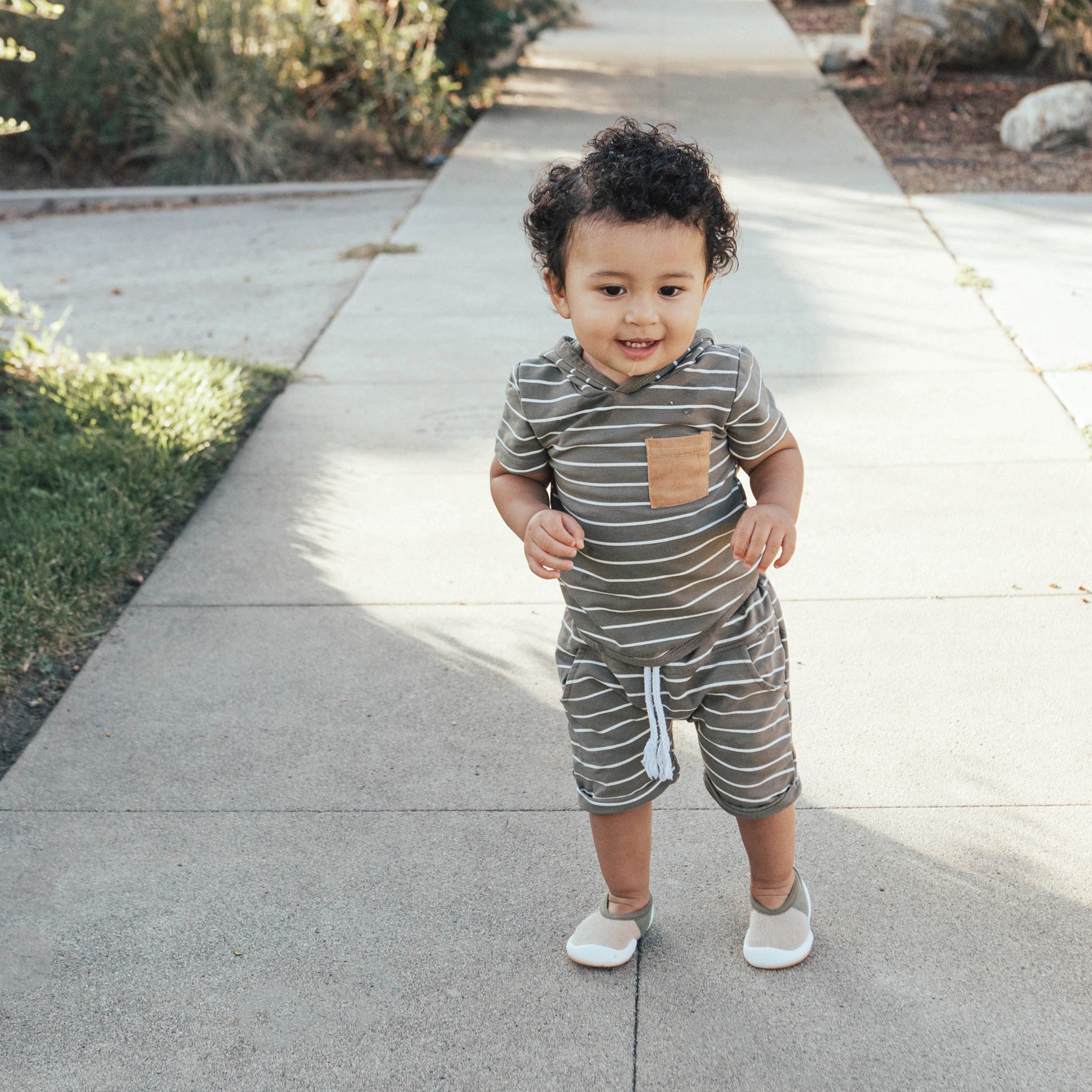 A toddler stands on a sidewalk wearing a striped short set and moccasin-style shoes.