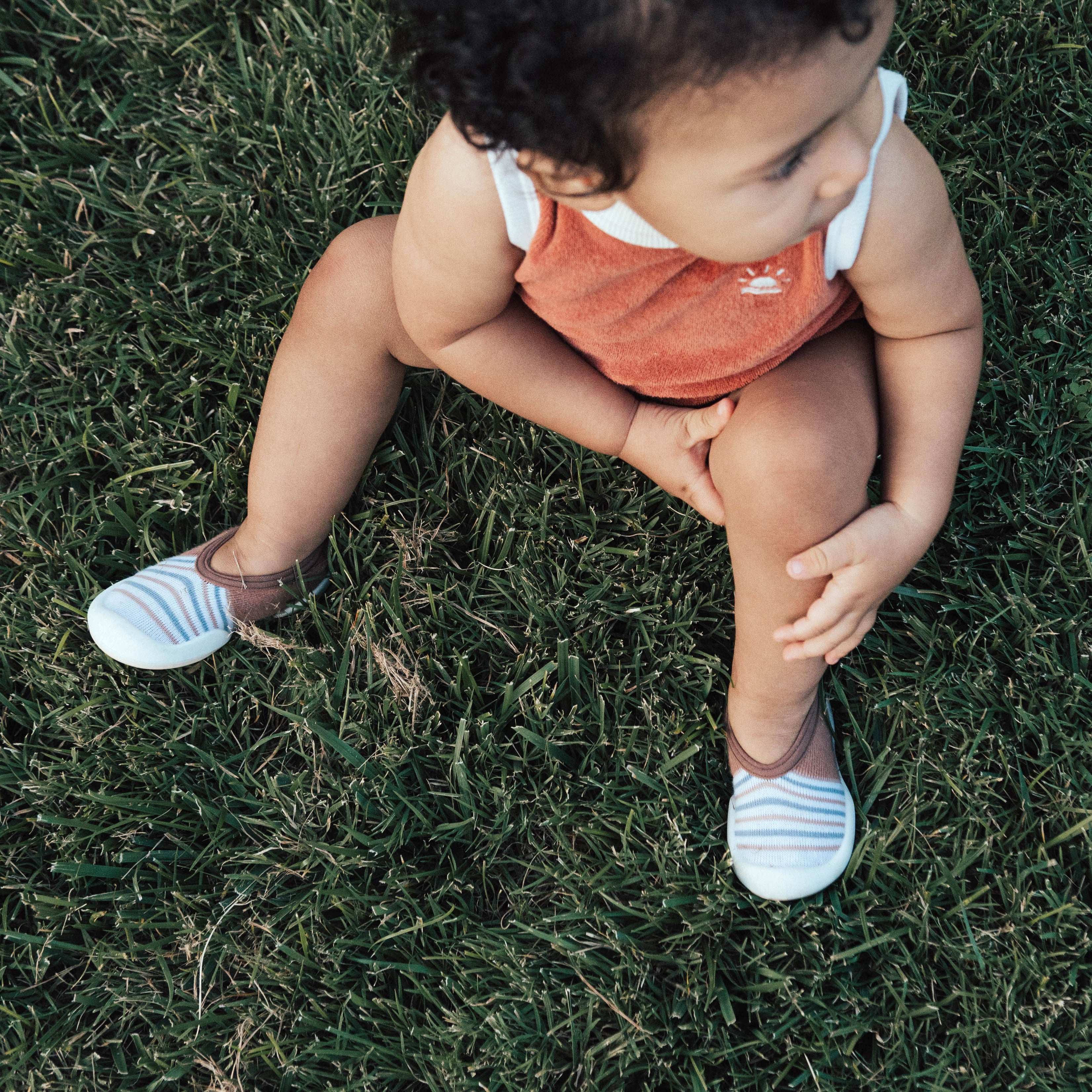 Baby sitting on grass wearing a red romper and striped shoes.