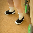 A toddler stands on a cork floor wearing black and white slipper shoes and beige ribbed socks, next to a toy walker.