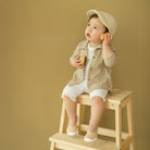A toddler wearing a cap and checked shirt sits on a wooden step stool and plays with wooden blocks.