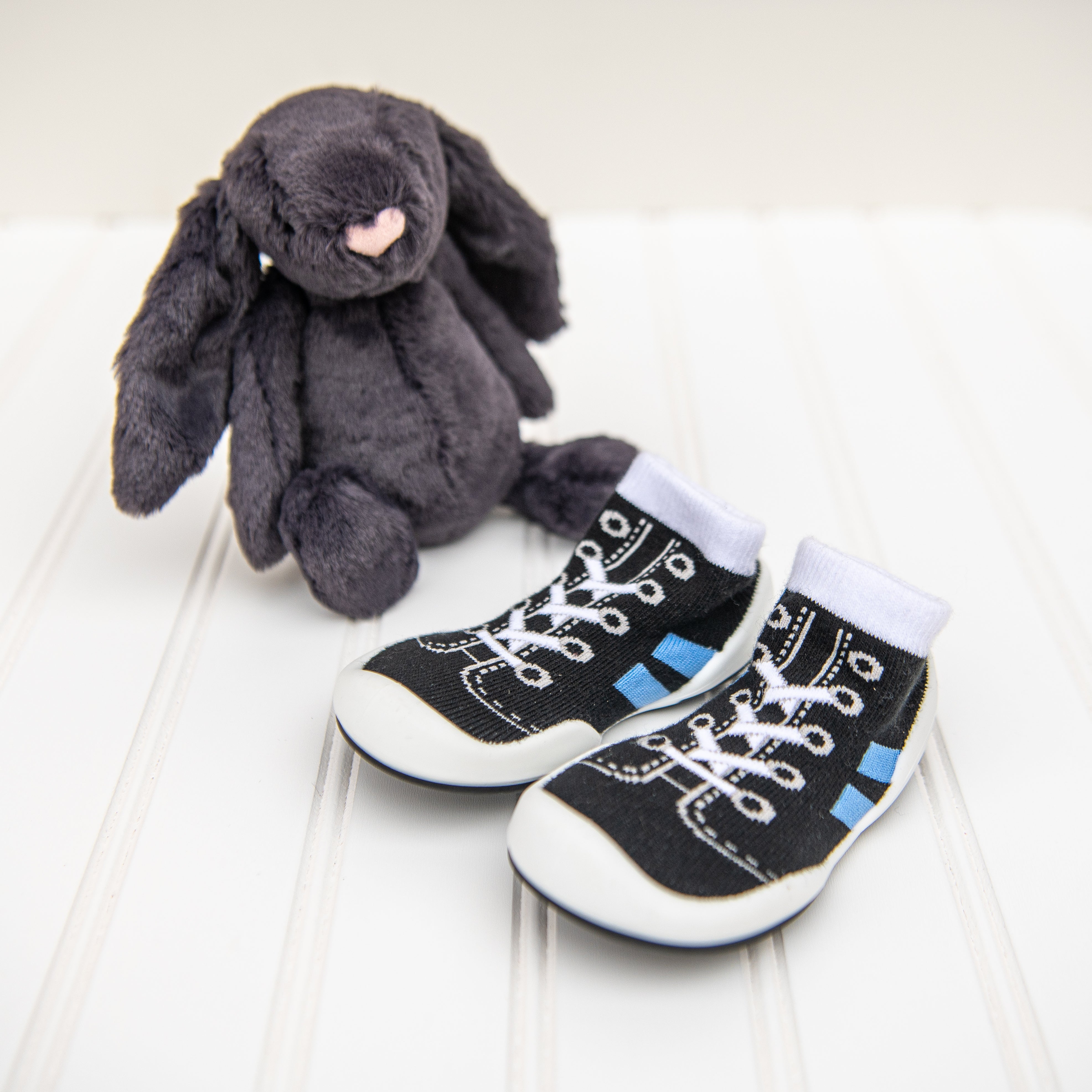 A grey bunny plush toy and a pair of black baby shoe socks with white soles.