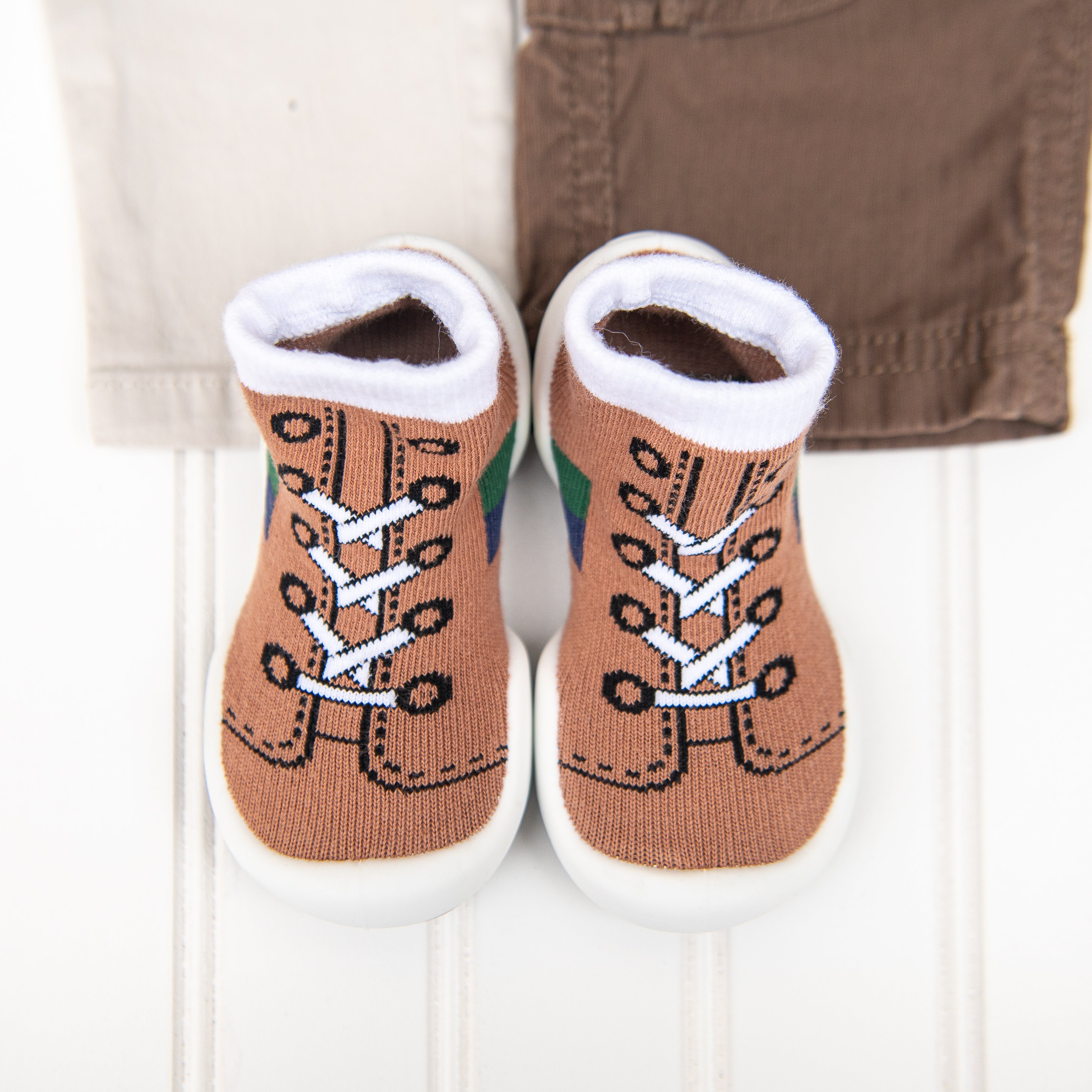 Brown and white baby socks shaped like shoes, with a pair of tan and brown pants in the background.