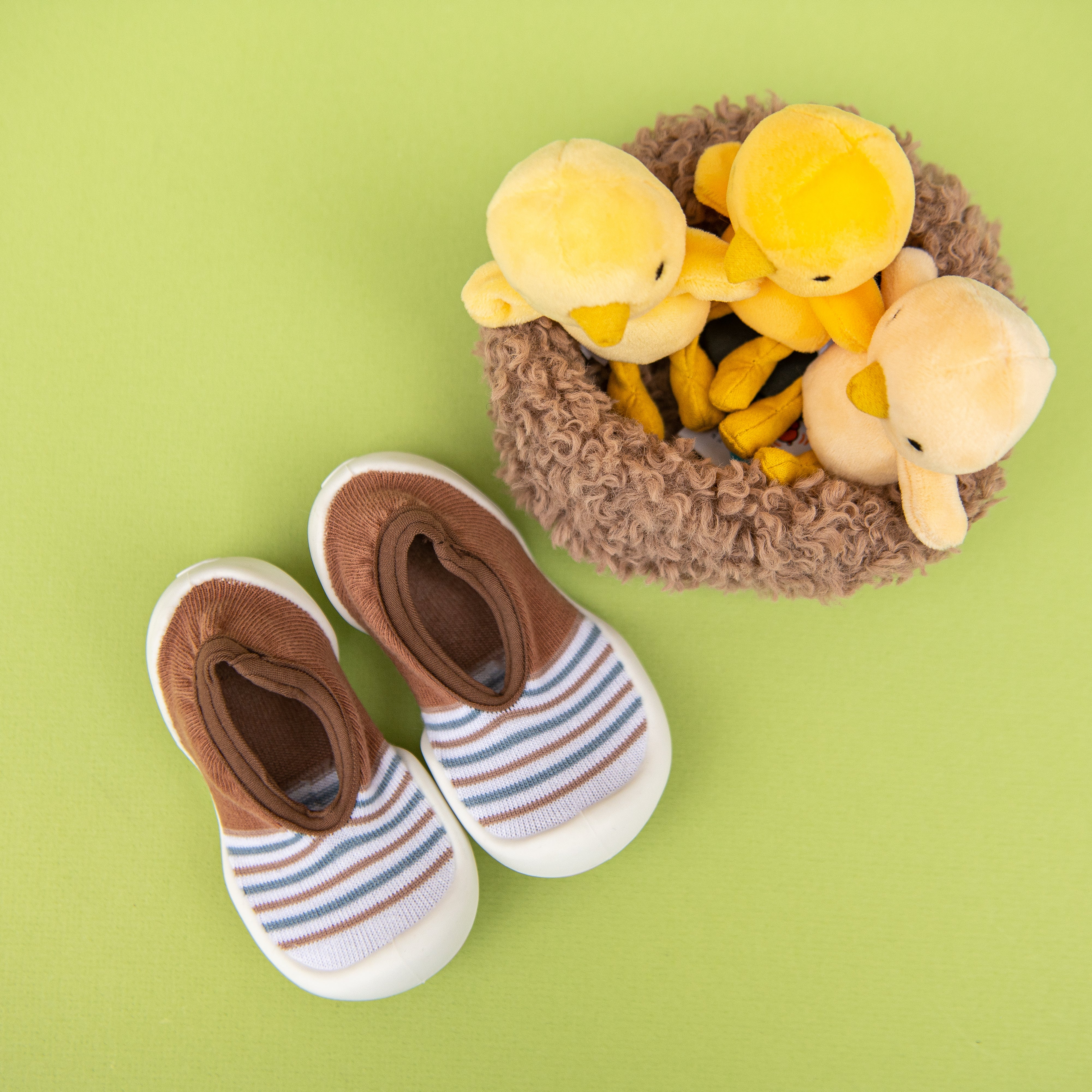 Top-down product shot: baby shoes, duckling toys, nest, green background.