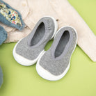 A pair of gray baby shoes with white soles sits on a green surface next to a light beige knit cardigan with floral-lined trim.