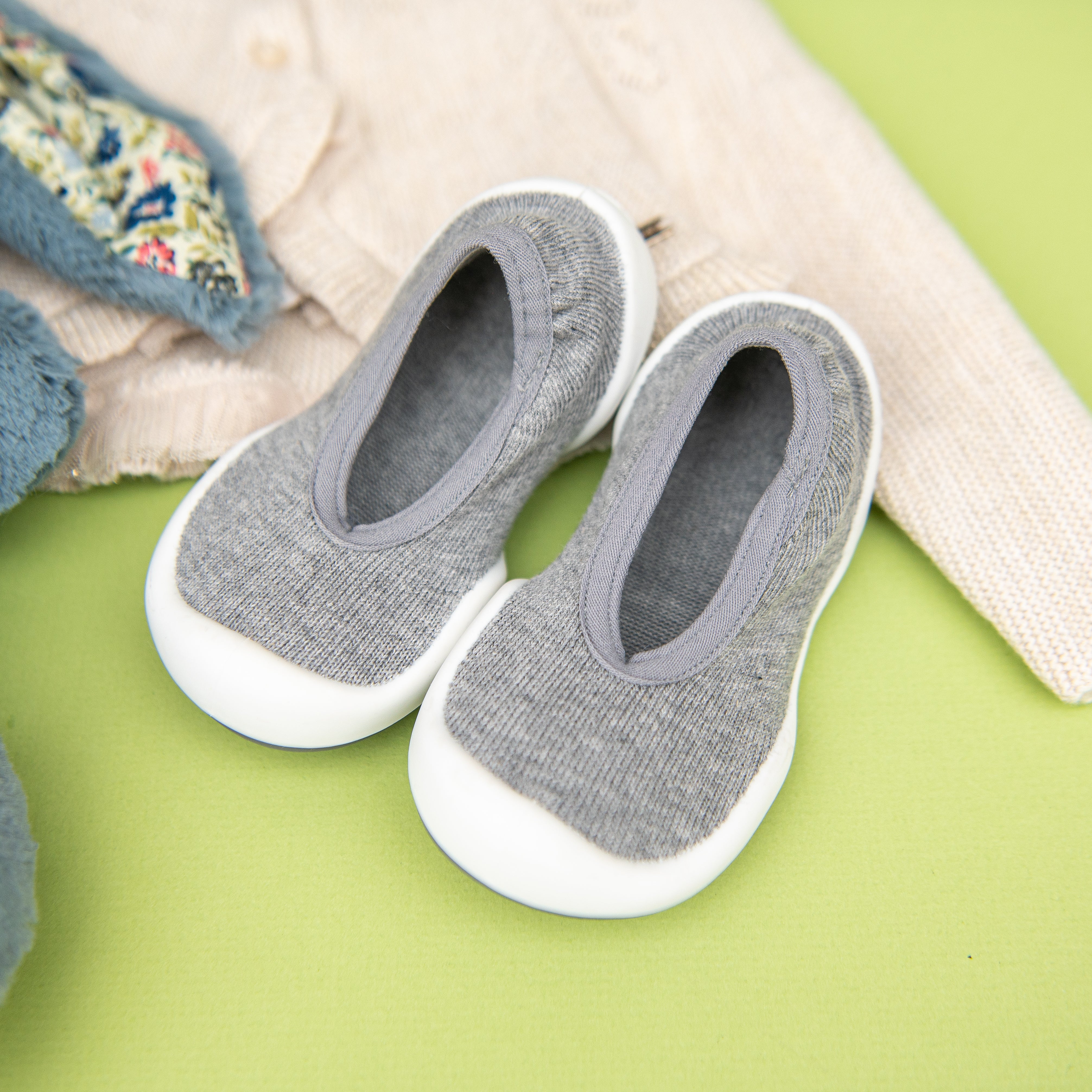 A pair of gray baby shoes with white soles sits on a green surface next to a light beige knit cardigan with floral-lined trim.
