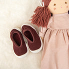 Doll with brown yarn hair and a pink dress next to a pair of brown and white baby shoes on a white, textured surface.