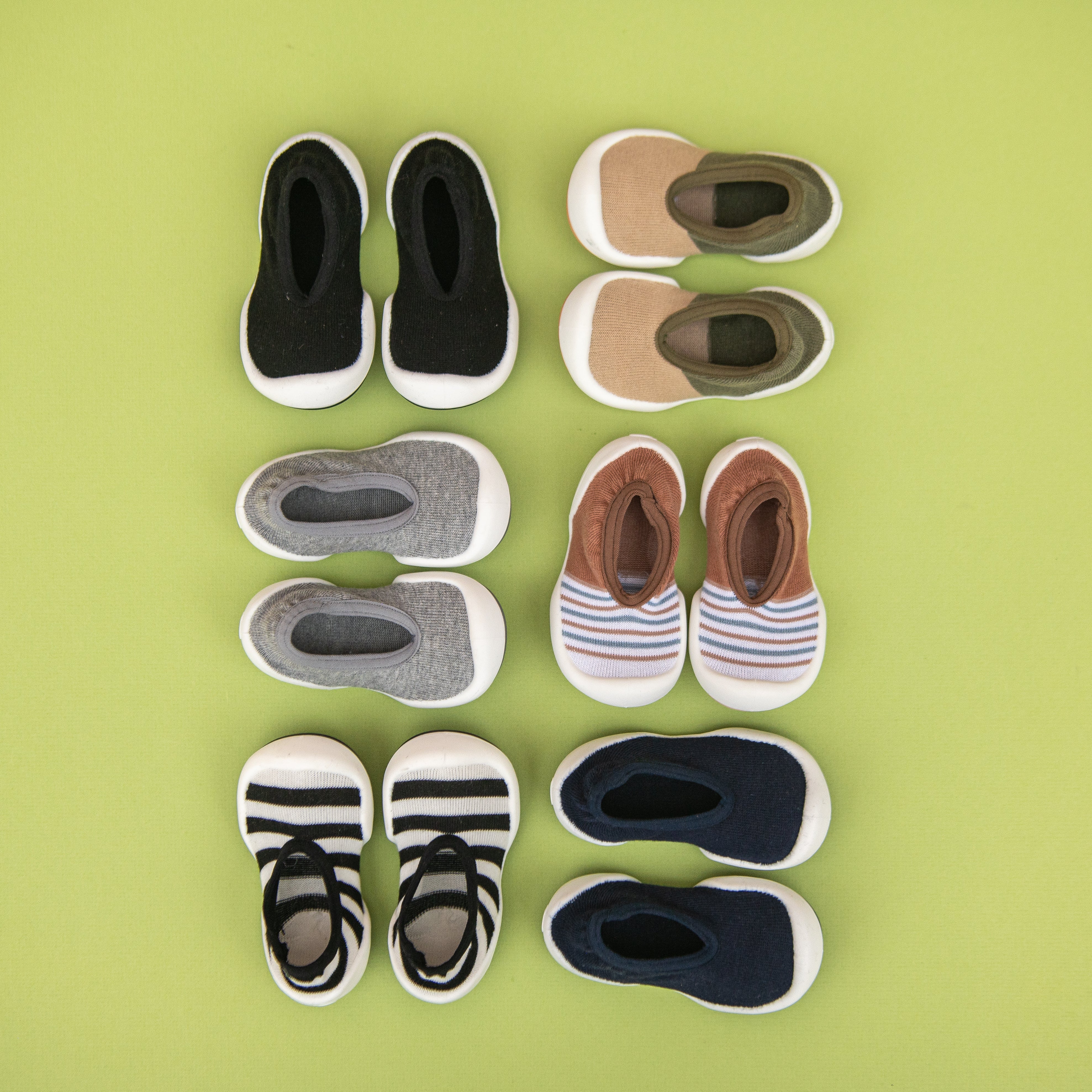 Six pairs of baby walking shoes in various colors, laid out on a light green background.