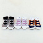 Three pairs of baby socks resembling sneakers. Gray, purple, and brown color schemes. White soles and soft fabric construction.