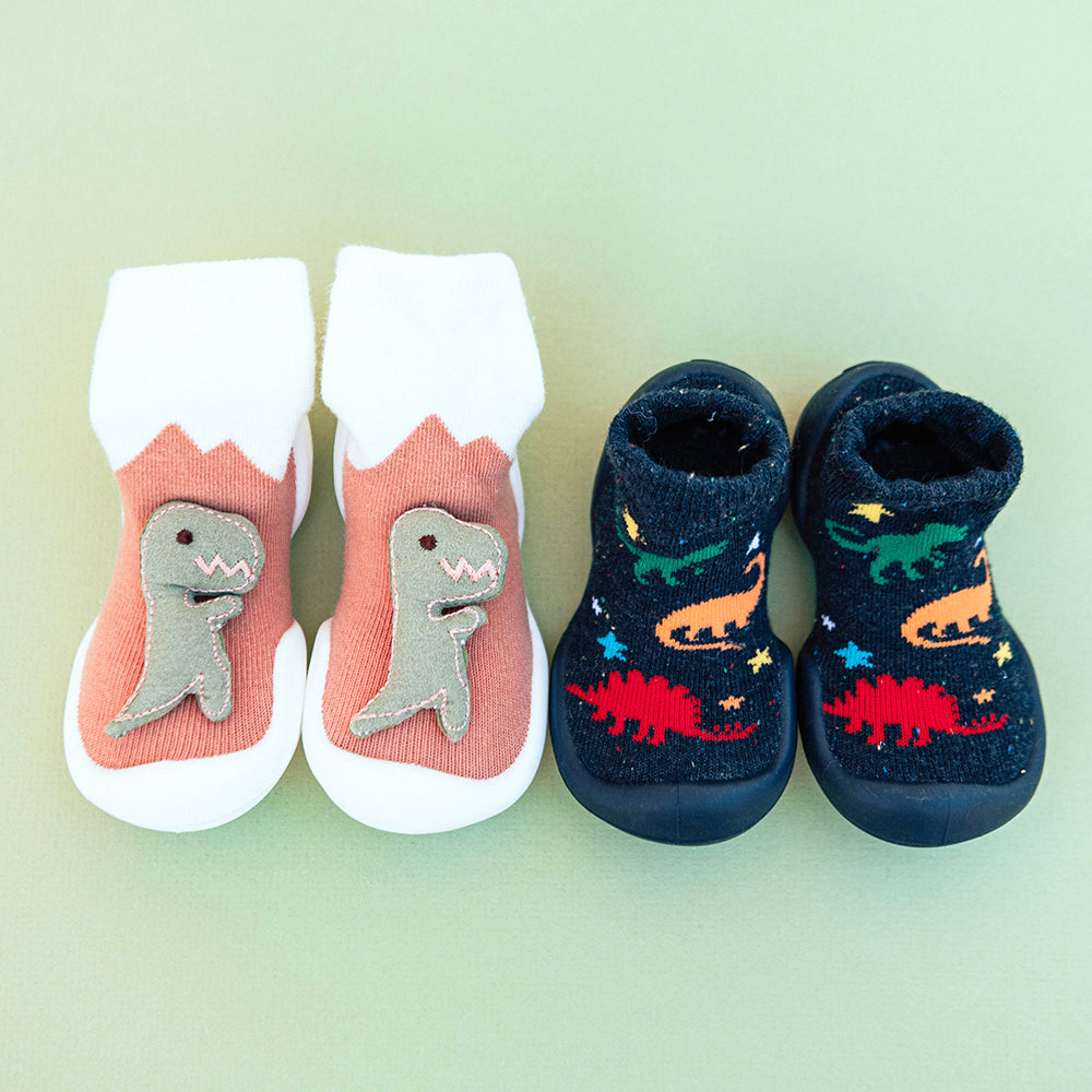 Two pairs of baby socks, one pair with dinosaurs on the front and a white cuff, and one dark pair with dinosaurs and stars printed on them.