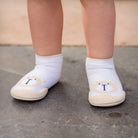 Toddler's legs and feet wearing white bear-themed Koemullo sock shoes with tan rubber soles on a gray surface.