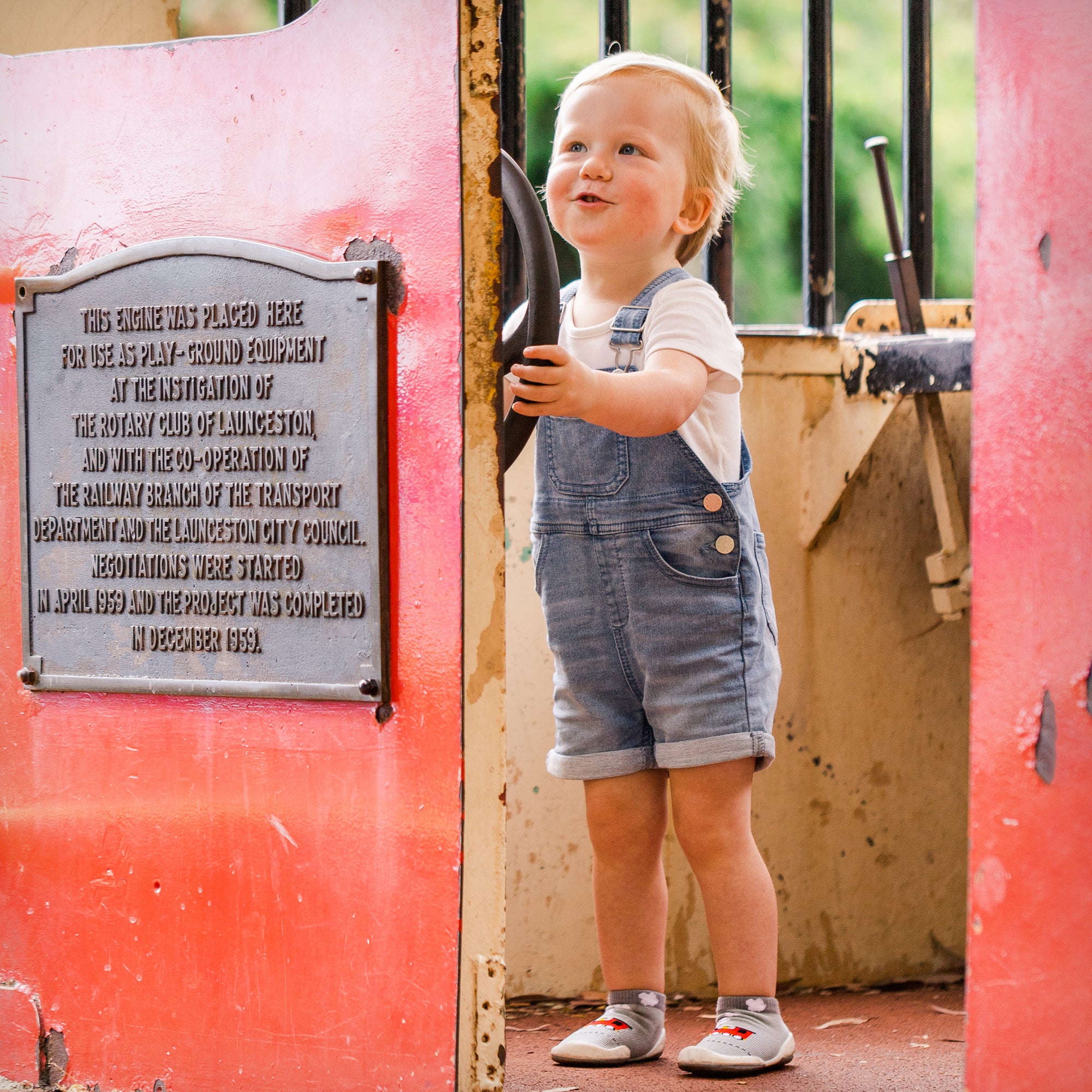 Young child in denim overalls and white shirt, holding a black handle, next to a red structure with a plaque.