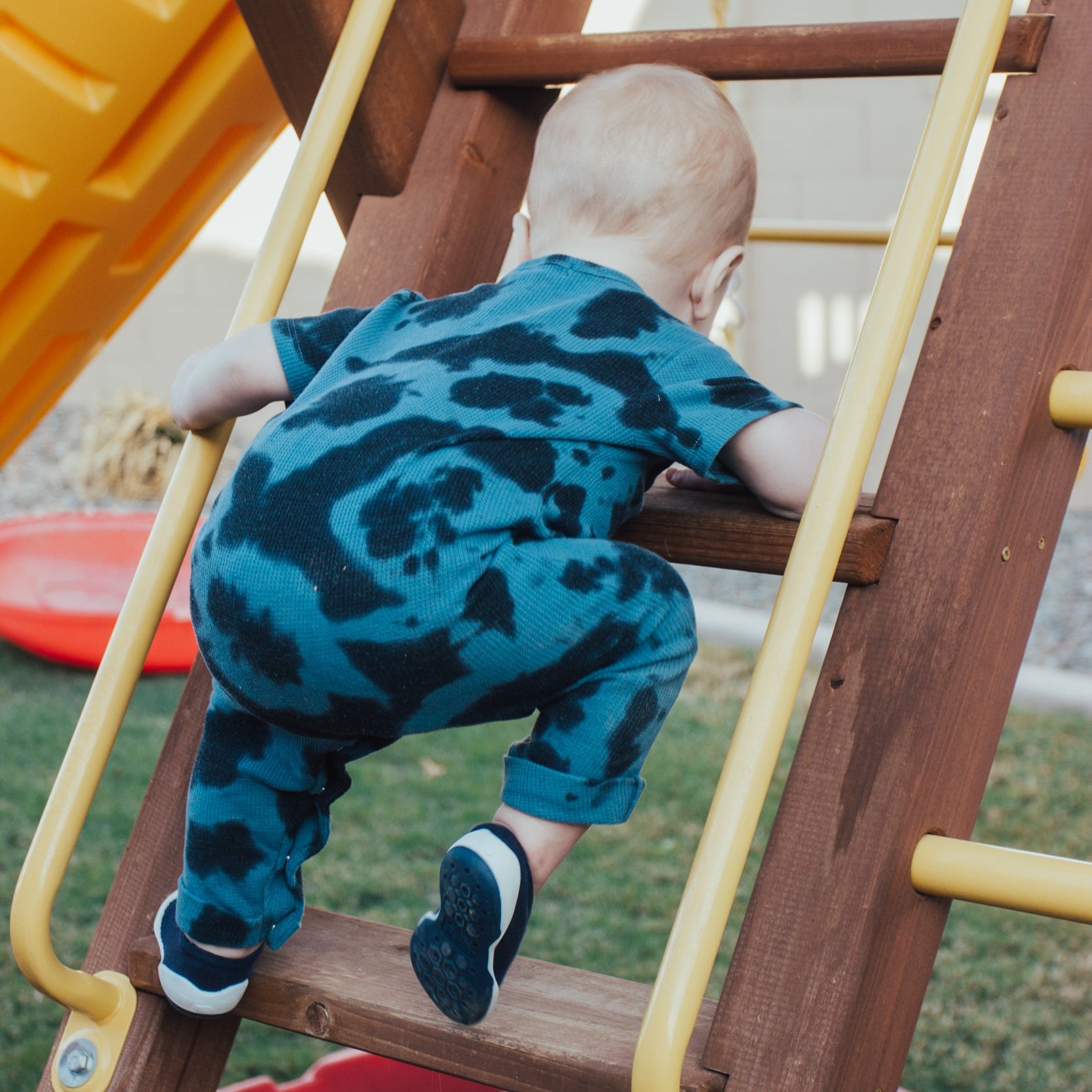 Child in blue tie-dye outfit climbs wooden ladder with yellow bars.