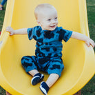 Baby boy in a blue tie-dye outfit sits on a yellow slide with arms outstretched.