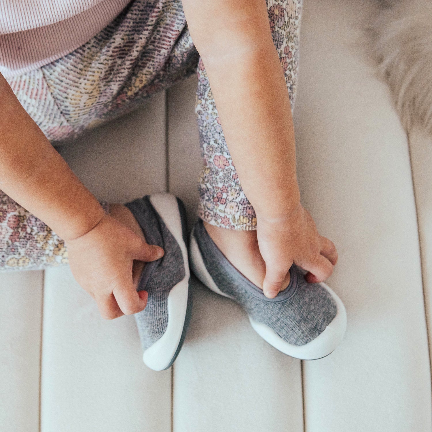 Child putting on gray and white shoes while wearing floral pants.