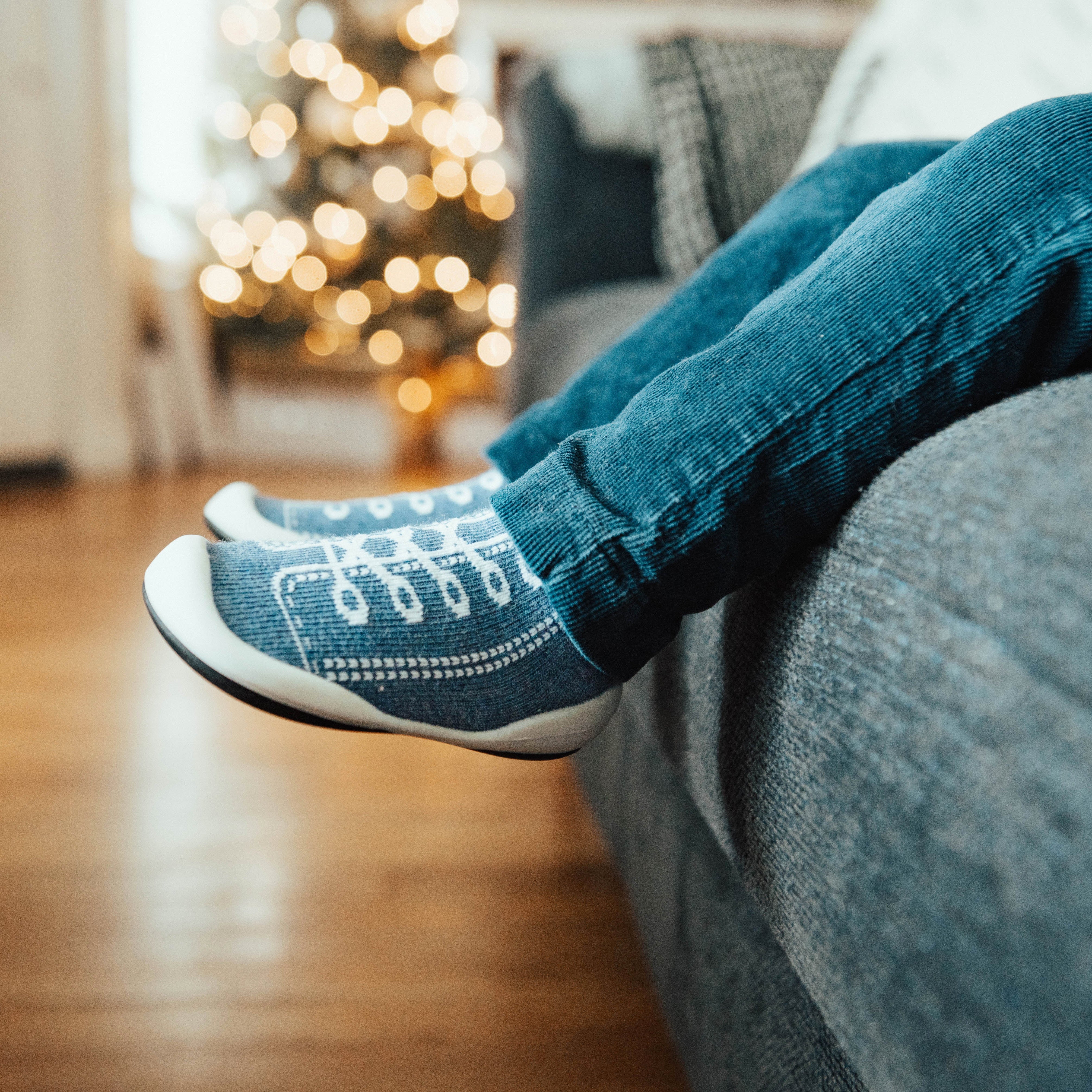 Person wearing blue corduroy pants and blue patterned socks, sitting on a gray couch. Christmas tree with lights in the background.