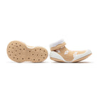 Two tan, sock-like toddler shoes with white flowers and white trim. One shoe displays the honeycomb-patterned sole.