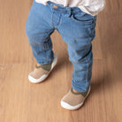 A toddler wearing light-wash denim jeans and beige and olive socks with white soles stands on a wooden floor.