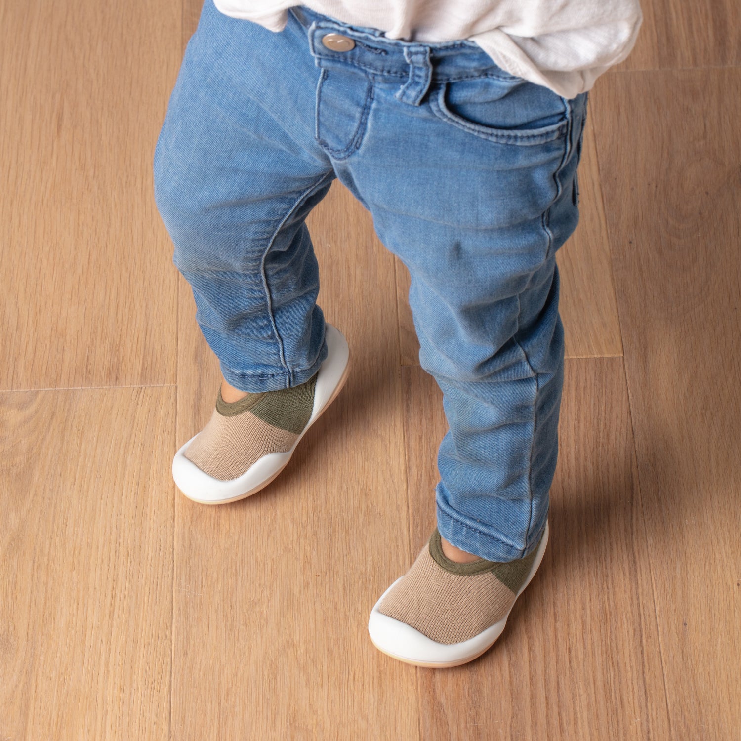 A toddler wearing light-wash denim jeans and beige and olive socks with white soles stands on a wooden floor.
