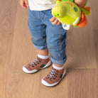 A toddler wears white socks and brown shoe-socks on a wooden floor, holding a plush green dinosaur.