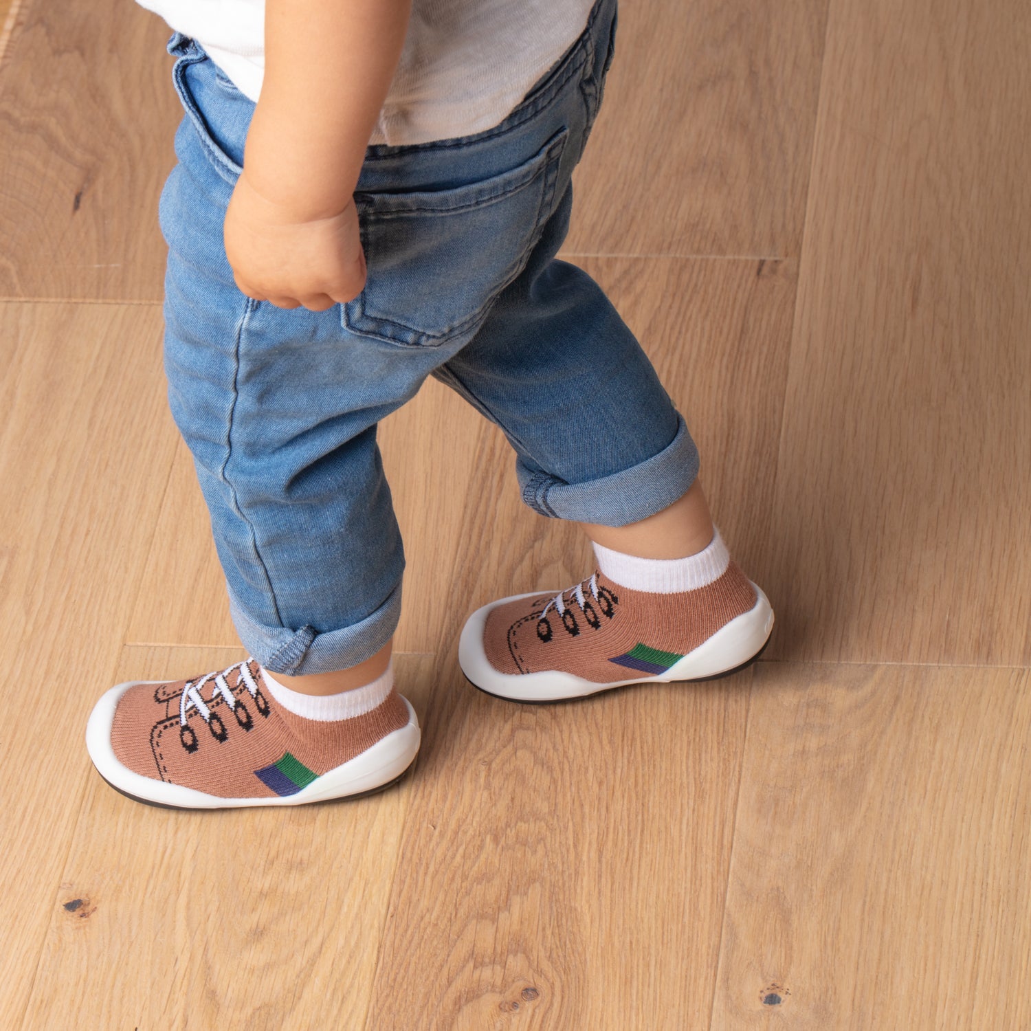 A child in jeans and sock-shoes stands on a wood floor.