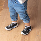 Child standing in blue jeans and black sneaker socks on a wood floor.