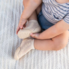 Toddler with sock-like beige shoes with rubber soles sitting on a patterned white blanket.