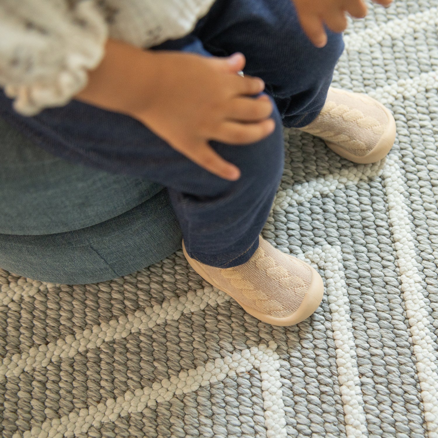 Child sitting, wearing blue pants and beige slipper-socks, on a gray and white rug.