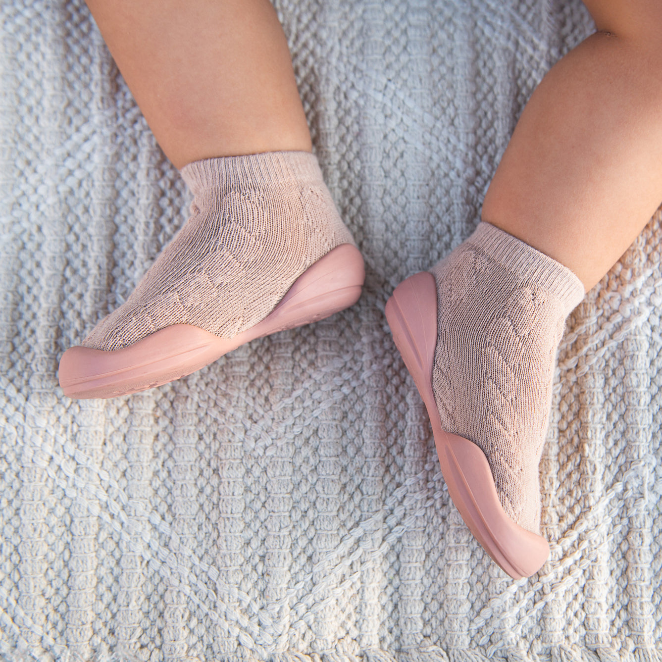 Overhead shot of a baby's feet wearing pink sock shoes on a patterned off-white blanket.
