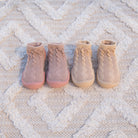 Four infant sock-shoes in shades of pink and beige arranged on a patterned white rug.