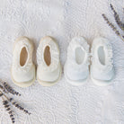 Two pairs of baby shoes, one pair off-white and one pair white, on a white textured fabric background with sprigs of lavender.