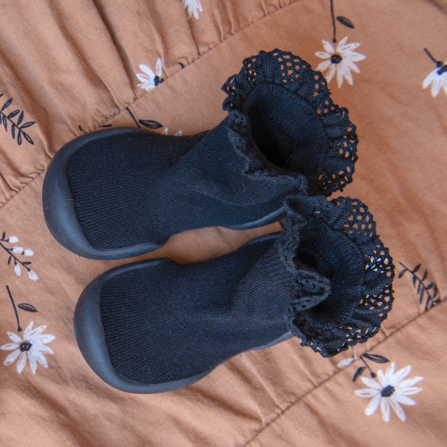 Black baby booties with lace on a brown floral fabric.