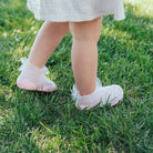 Close-up of a baby's legs and feet in pink socks with white ruffles, standing on green grass.