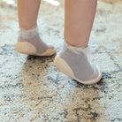 Close-up of a baby's feet in grey sock shoes with white lace trim on a textured rug.