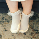Baby's legs and feet in white socks with lace trim and beige soles, standing on a patterned carpet.