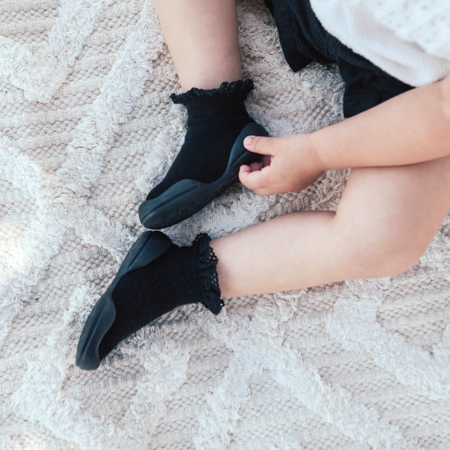 Child in black socks and shoes sitting on a white textured rug.