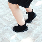 Close-up of a baby's legs in black socks and a black dress, standing on a light-colored textured rug.