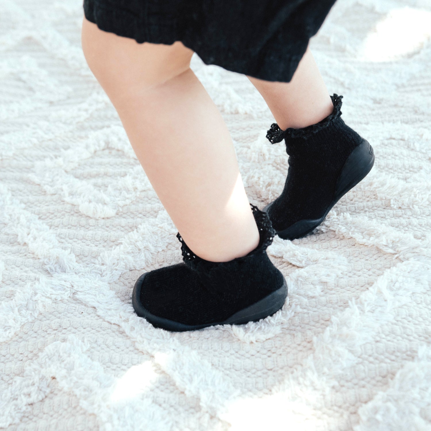 Close-up of a baby's legs in black socks and a black dress, standing on a light-colored textured rug.