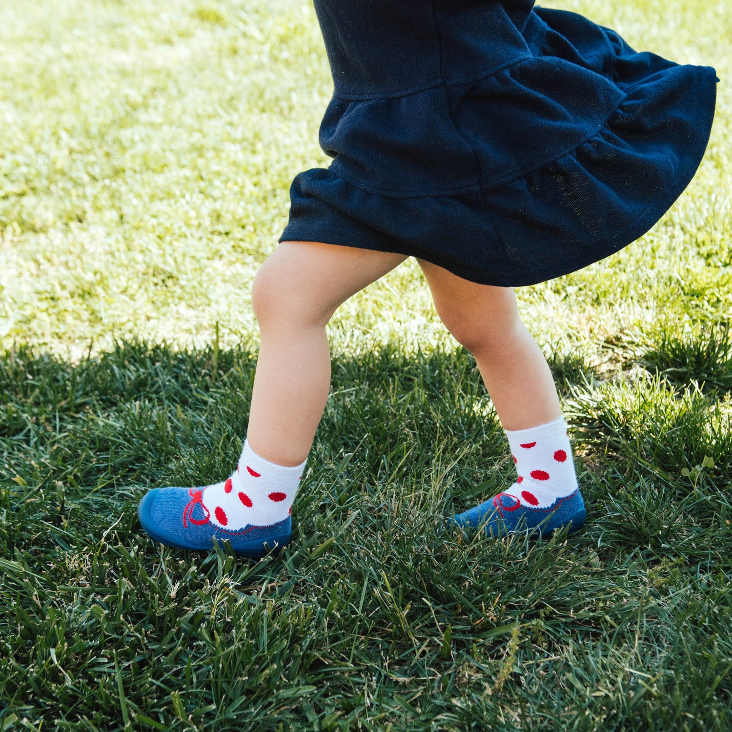 Child in blue dress with red-dotted socks and blue shoes on grass.