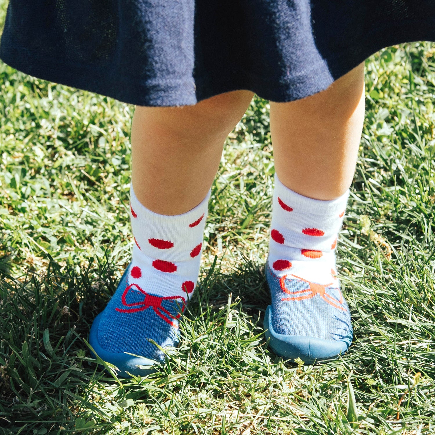 A child standing on grass wearing a blue dress, white socks with red dots, and blue shoes with red bows.