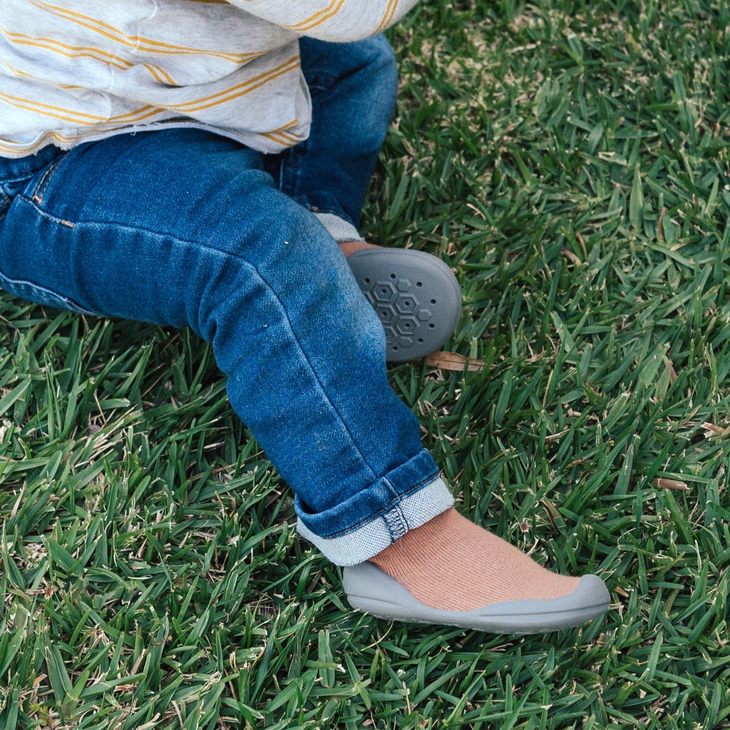 Child seated on grass wearing jeans, striped shirt, brown socks, and grey shoes.