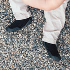 A person wearing tan pants and black minimalist shoes standing on gravel.