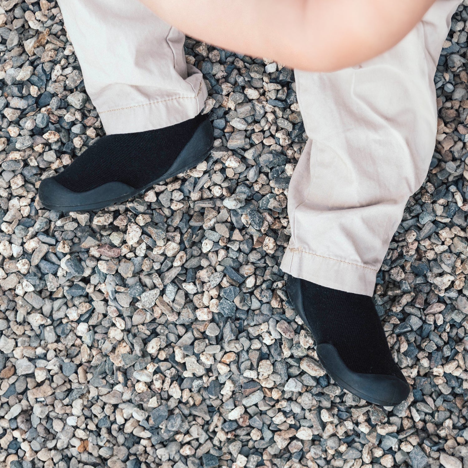 A person wearing tan pants and black minimalist shoes standing on gravel.