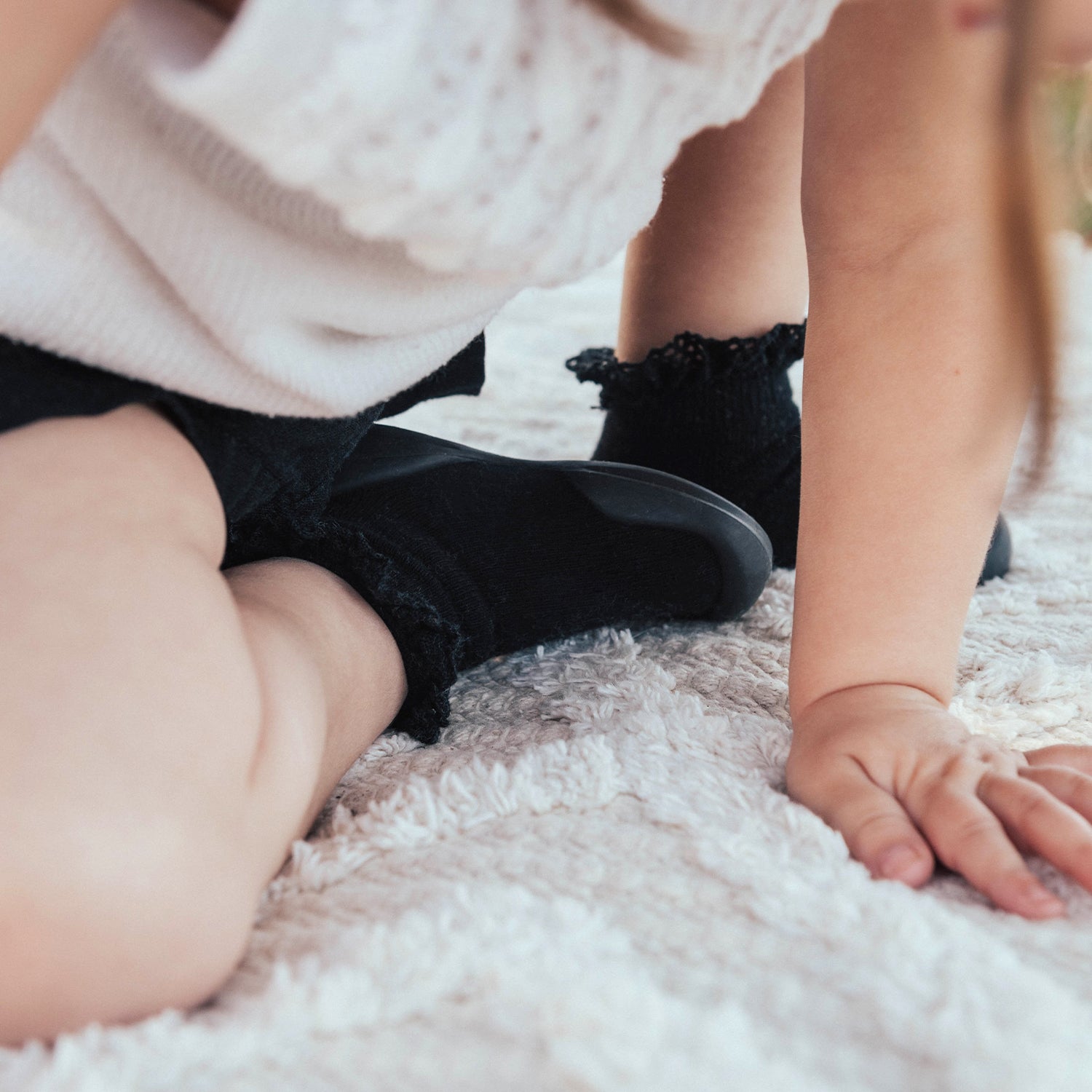 Child on white shag rug, wearing a white sweater, black shorts, and black socks.