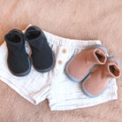 Two pairs of baby shoes, black and brown, on top of white shorts with wooden buttons.